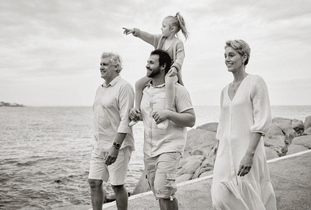 Three generations of a family walk together along a waterfront path, a young girl pointing from her father’s shoulders