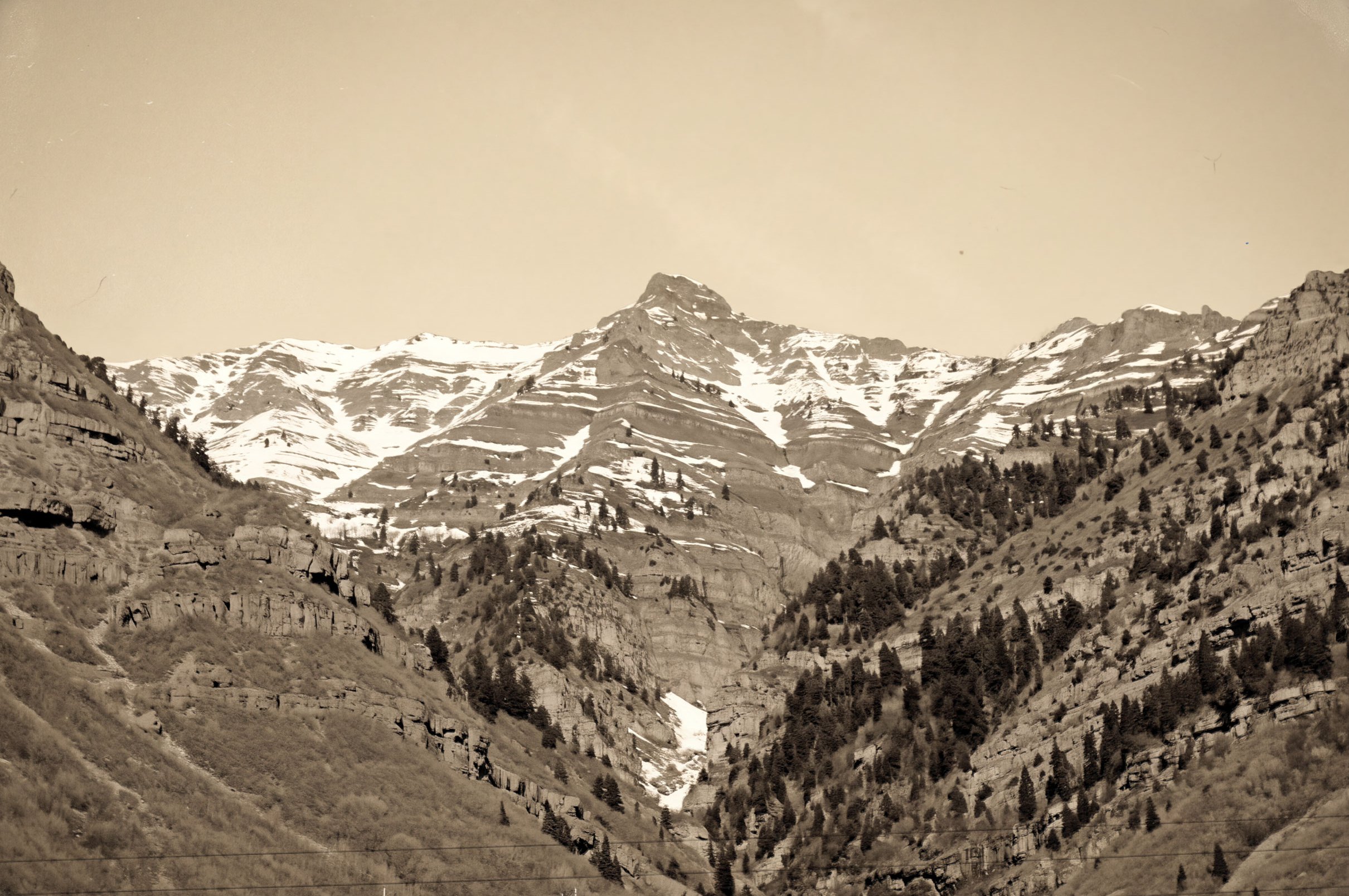 A sepia photograph of snow-streaked peaks rising above a steep, forested canyon